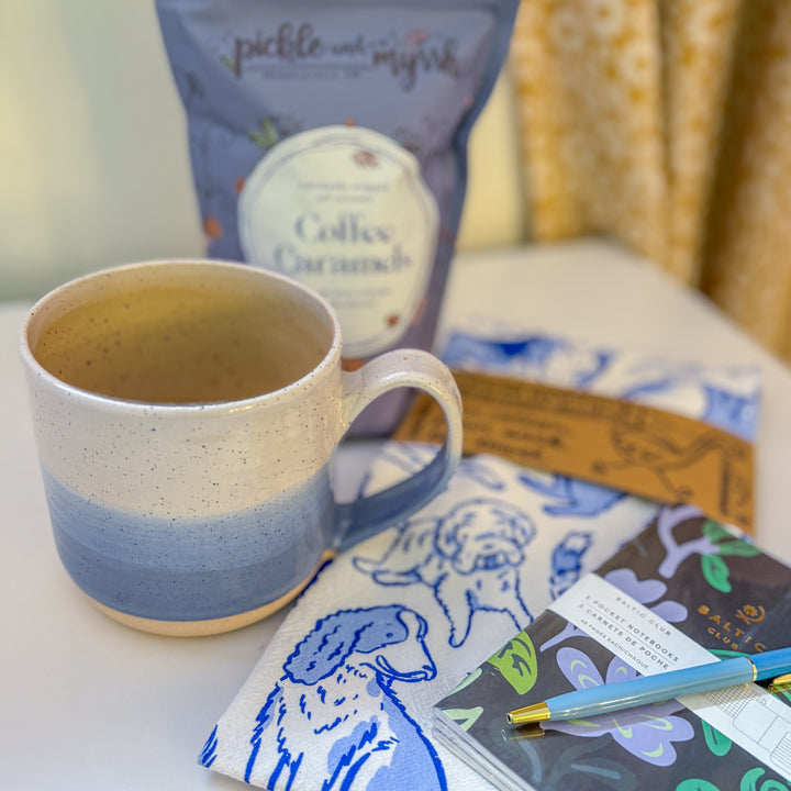 A handmade ceramic mug with a blue ombre glaze, featuring a speckled texture, displayed on a light gray background. The mug has a large, rounded handle and is made in Mississippi Mills, Ontario, Canada.