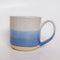 A handmade ceramic mug with a blue ombre glaze, featuring a speckled texture, displayed on a light gray background. The mug has a large, rounded handle and is made in Mississippi Mills, Ontario, Canada.