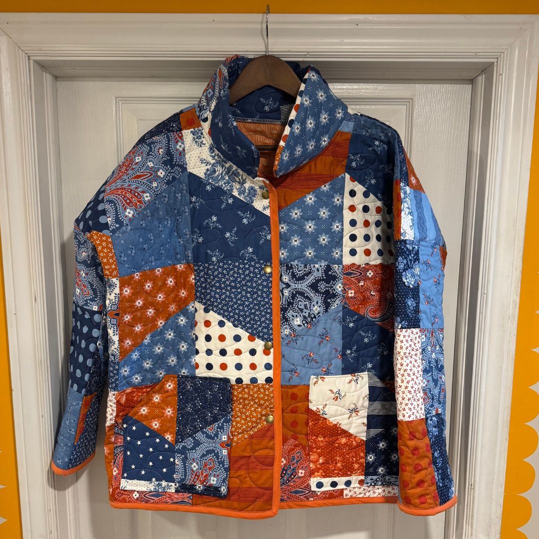 A quilted patchwork jacket hanging on a wooden hanger against a white paneled door, featuring a mix of blue, orange, and white patterned fabric panels.