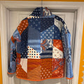 A handmade patchwork quilted jacket with long sleeves and a stand-up collar, hanging on a hanger in front of a white door. The jacket features a variety of blue, orange, and white patterned fabric panels.