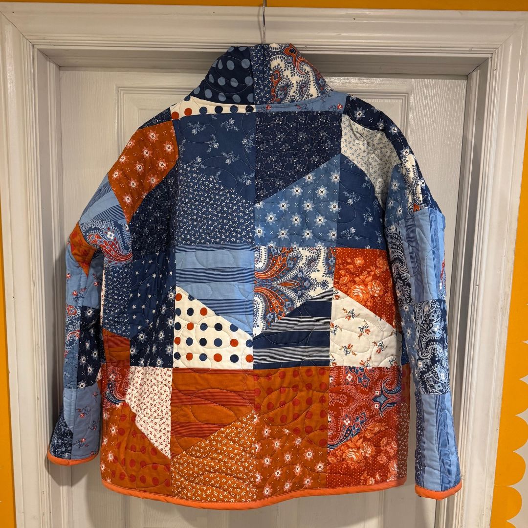 A handmade patchwork quilted jacket with long sleeves and a stand-up collar, hanging on a hanger in front of a white door. The jacket features a variety of blue, orange, and white patterned fabric panels.