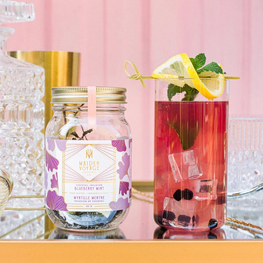 A glass jar filled with dried blueberries, mint leaves, and other ingredients. The jar has a white label with the Maiden Voyage logo and the product name "Blueberry Mint Cocktail Infusion."