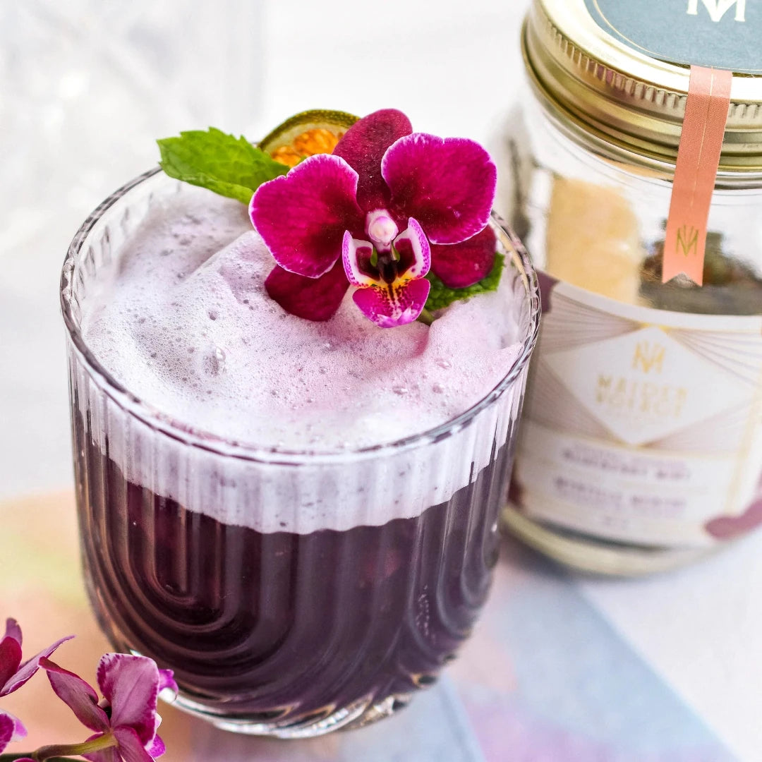 A glass jar filled with dried blueberries, mint leaves, and other ingredients. The jar has a white label with the Maiden Voyage logo and the product name "Blueberry Mint Cocktail Infusion."