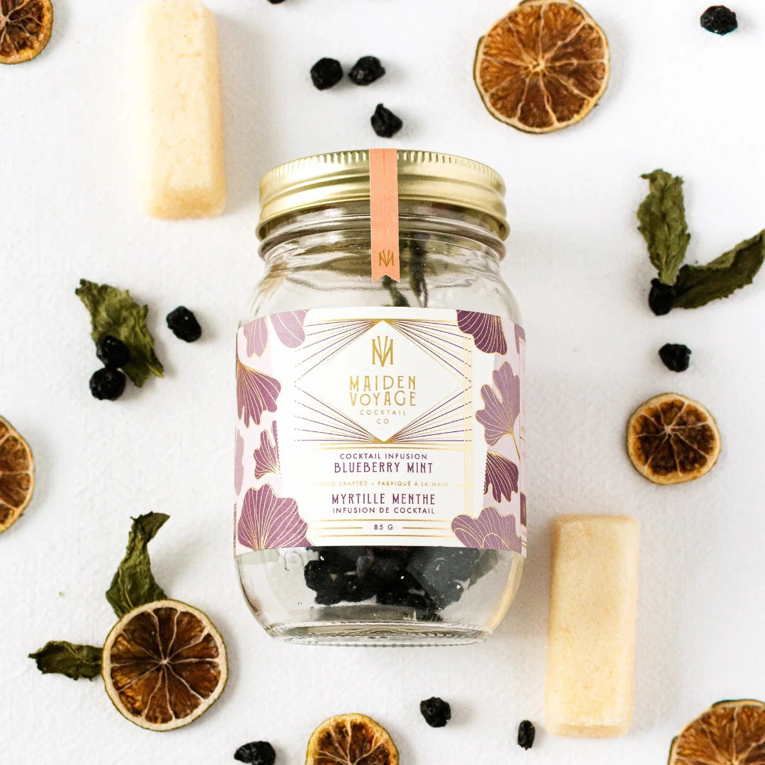 A glass jar filled with dried blueberries, mint leaves, and other ingredients. The jar has a white label with the Maiden Voyage logo and the product name "Blueberry Mint Cocktail Infusion."