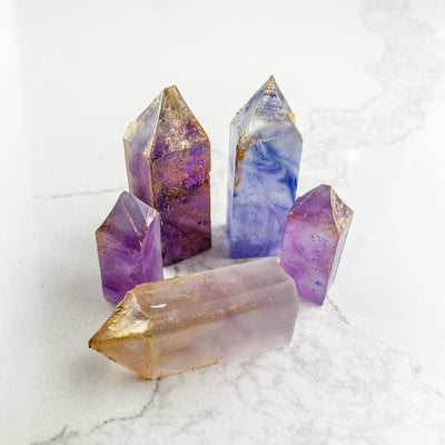 A collection of purple, crystal-shaped candies arranged on a white marble surface. Some candies have gold accents. The candies have a crystal-like appearance with a slightly translucent quality.