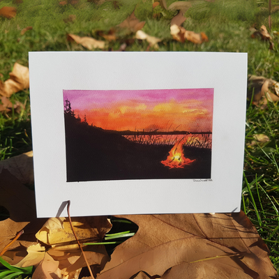 A watercolor greeting card of a campfire on a beach at sunset. The sky is ablaze with orange and pink hues, reflecting on the calm water. Silhouettes of trees and hills frame the scene, with a small campfire casting a warm glow on the beach in the foreground.