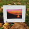 A watercolor greeting card of a campfire on a beach at sunset. The sky is ablaze with orange and pink hues, reflecting on the calm water. Silhouettes of trees and hills frame the scene, with a small campfire casting a warm glow on the beach in the foreground.