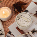 Small candle and decorative container on a wooden surface with a lit candle and leaves.