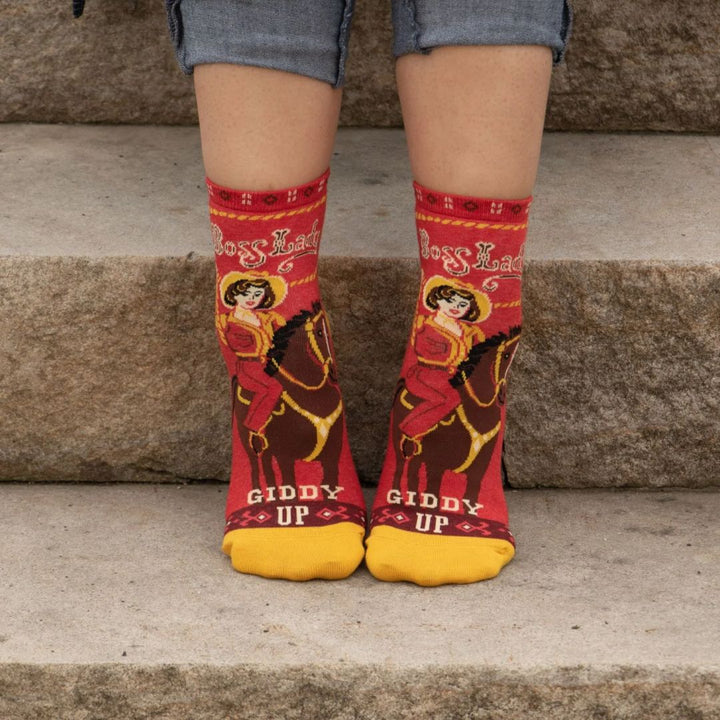 A pair of red ankle socks with yellow accents. One sock features a vintage-style illustration of a cowgirl riding a horse, with 