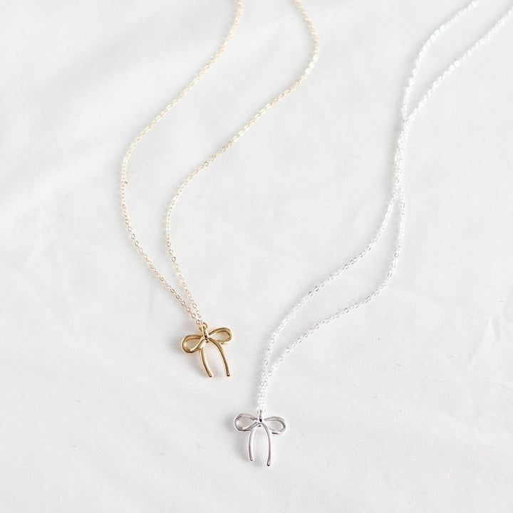 Two necklaces on a white plate. One necklace has a gold bow pendant, and the other has a silver bow pendant.