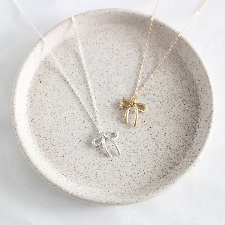Two necklaces on a white plate. One necklace has a gold bow pendant, and the other has a silver bow pendant.