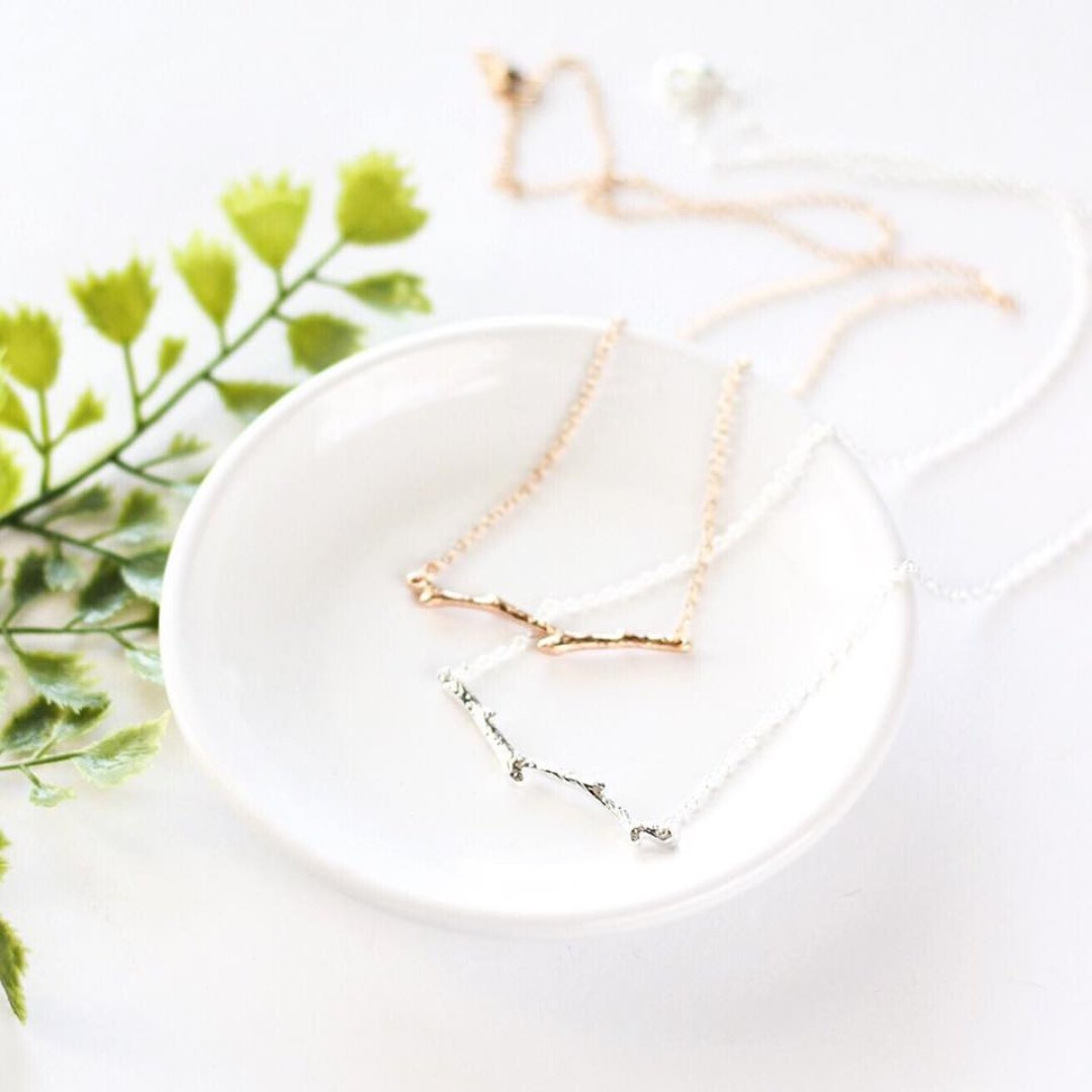 Two necklaces with branch pendants on a white surface. One necklace has a gold branch pendant, and the other has a silver branch pendant.
