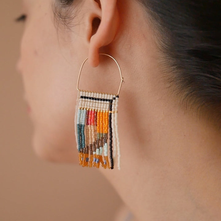 A pair of rectangular, tapestry-style earrings hand-stitched with small Japanese glass beads in a geometric pattern featuring shades of cream, orange, pink, light blue, teal, brown, and black. They hang from delicate, half-circle 14 KT gold filled wire hoops.