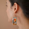 A pair of rectangular, tapestry-style earrings hand-stitched with small Japanese glass beads in a geometric pattern featuring shades of cream, orange, pink, light blue, teal, brown, and black. They hang from delicate, half-circle 14 KT gold filled wire hoops.