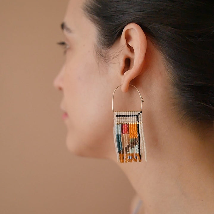 A pair of rectangular, tapestry-style earrings hand-stitched with small Japanese glass beads in a geometric pattern featuring shades of cream, orange, pink, light blue, teal, brown, and black. They hang from delicate, half-circle 14 KT gold filled wire hoops.