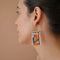 A pair of rectangular, tapestry-style earrings hand-stitched with small Japanese glass beads in a geometric pattern featuring shades of cream, orange, pink, light blue, teal, brown, and black. They hang from delicate, half-circle 14 KT gold filled wire hoops.