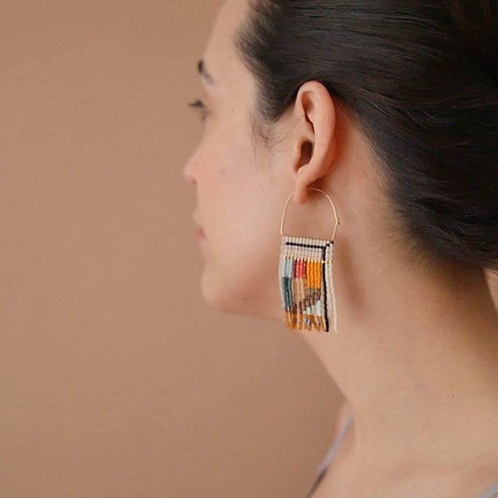 A pair of rectangular, tapestry-style earrings hand-stitched with small Japanese glass beads in a geometric pattern featuring shades of cream, orange, pink, light blue, teal, brown, and black. They hang from delicate, half-circle 14 KT gold filled wire hoops.