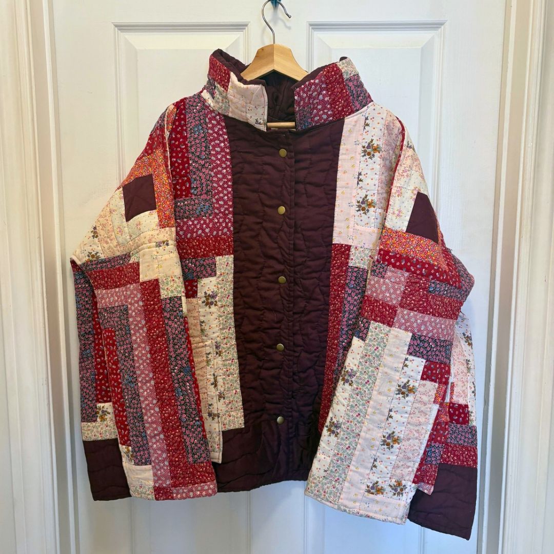 A handmade quilted jacket with a dark brown center panel and patchwork quilted sleeves and collar in red, beige, and floral patterns, hanging on a wooden hanger against a white paneled door.