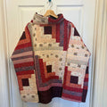A handmade quilted patchwork jacket with a mock neck, hanging on a wooden hanger against a white paneled door. The jacket features a geometric log cabin pattern in shades of red, brown, beige, and gray.