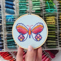 An overhead shot of a finished cross-stitch piece in a wooden embroidery hoop, displayed on a white surface with a potted succulent in the top right corner. The cross-stitch design features a colourful butterfly with wings in shades of orange, pink, and red, and blue accents, against a light blue background.