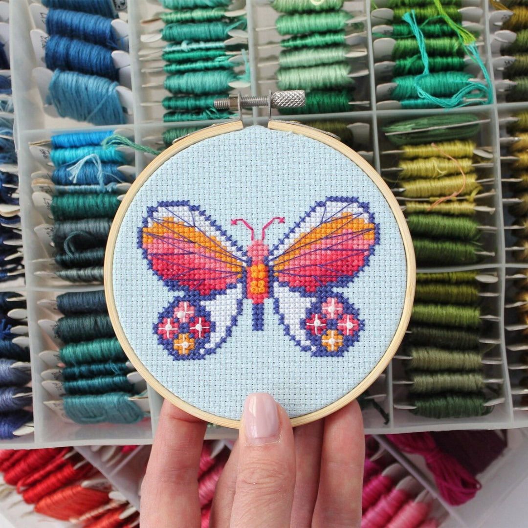An overhead shot of a finished cross-stitch piece in a wooden embroidery hoop, displayed on a white surface with a potted succulent in the top right corner. The cross-stitch design features a colourful butterfly with wings in shades of orange, pink, and red, and blue accents, against a light blue background.