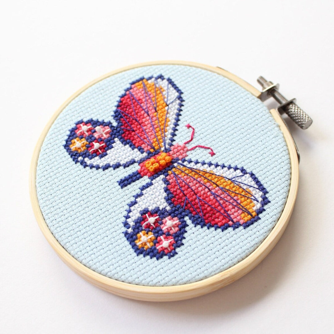 An overhead shot of a finished cross-stitch piece in a wooden embroidery hoop, displayed on a white surface with a potted succulent in the top right corner. The cross-stitch design features a colourful butterfly with wings in shades of orange, pink, and red, and blue accents, against a light blue background.