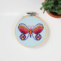 An overhead shot of a finished cross-stitch piece in a wooden embroidery hoop, displayed on a white surface with a potted succulent in the top right corner. The cross-stitch design features a colourful butterfly with wings in shades of orange, pink, and red, and blue accents, against a light blue background.