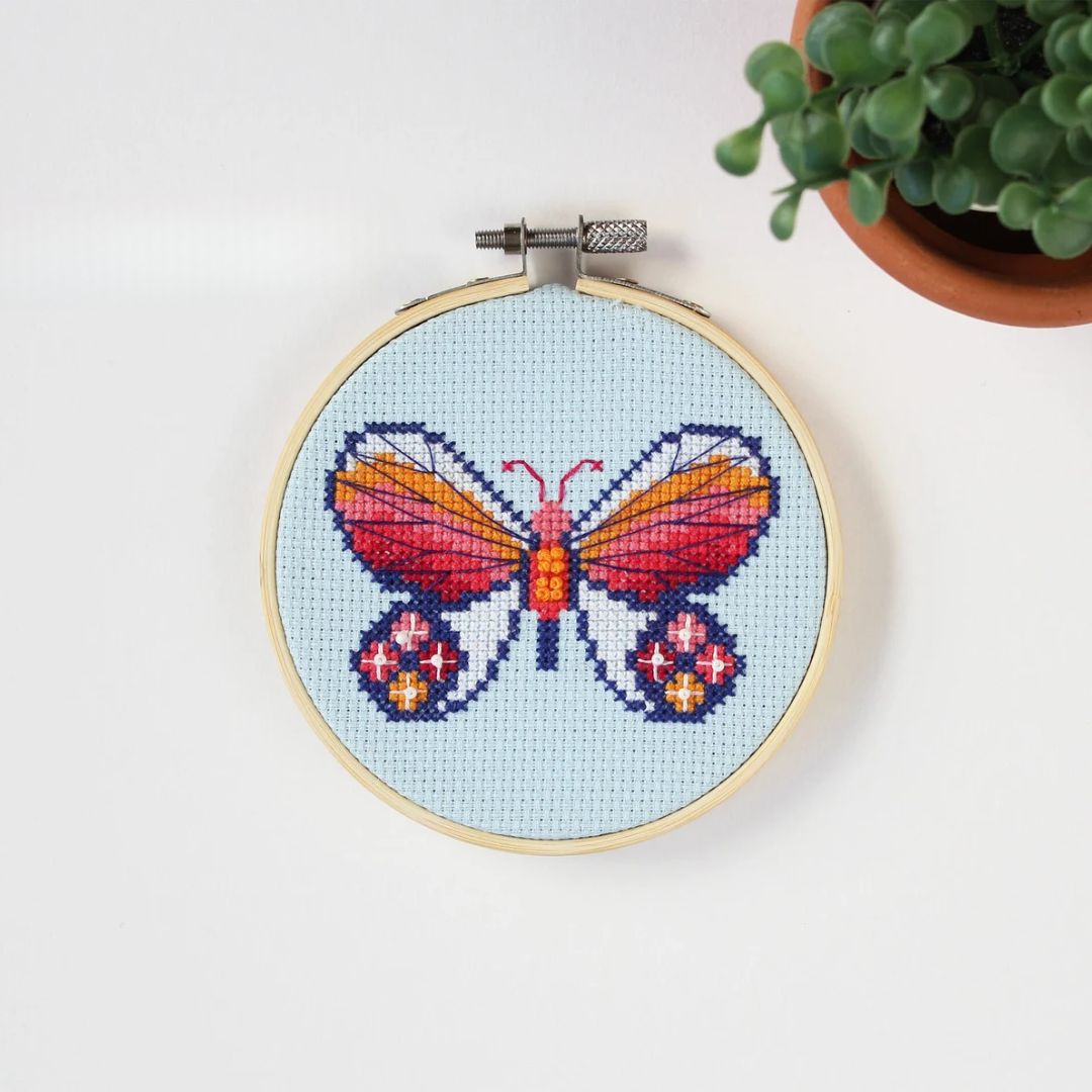 An overhead shot of a finished cross-stitch piece in a wooden embroidery hoop, displayed on a white surface with a potted succulent in the top right corner. The cross-stitch design features a colourful butterfly with wings in shades of orange, pink, and red, and blue accents, against a light blue background.