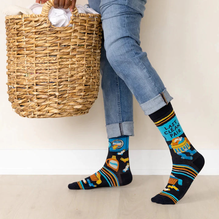 A pair of black socks with a laundry-themed design. The socks feature a washing machine, detergent bottles, a laundry basket, and socks themselves. It reads 