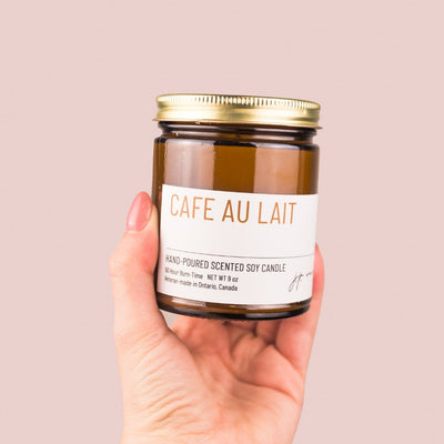 Hand holding a candle labeled 'Cafe Au Lait' against a pink background