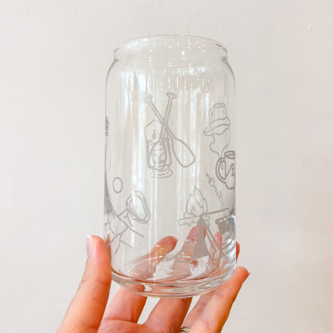 A hand holding a clear glass can-shaped tumbler with etched camping-themed illustrations, including a lantern, canoe, campfire, and hat, against a plain off-white wall.