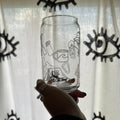 A hand holding a clear glass tumbler with black line art illustrations, set against a white curtain with black eye-like patterns.
