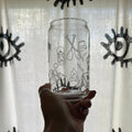 A hand holding a clear glass tumbler with black line-art illustrations of camping and outdoor gear, set against a white curtain with black eye-like patterns.