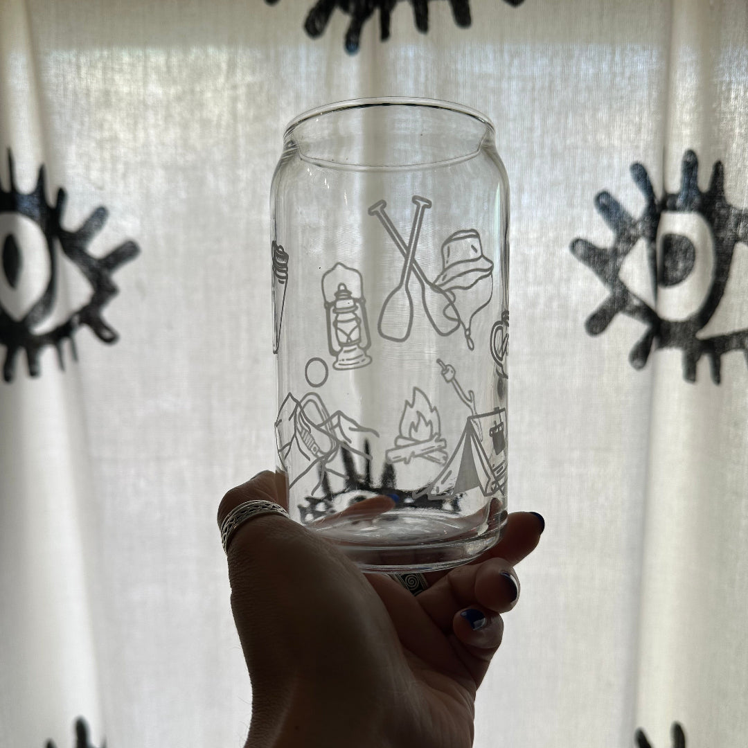 A hand holding a clear glass tumbler with black line-art illustrations of camping and outdoor gear, set against a white curtain with black eye-like patterns.