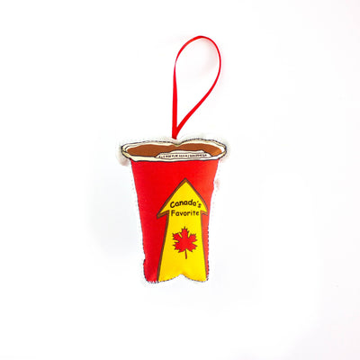 A close-up, high-angle flat lay shot on a plain white background features a soft, plush Christmas ornament shaped like a disposable coffee cup. The cup is red with a yellow and red arrow graphic on the front that says 