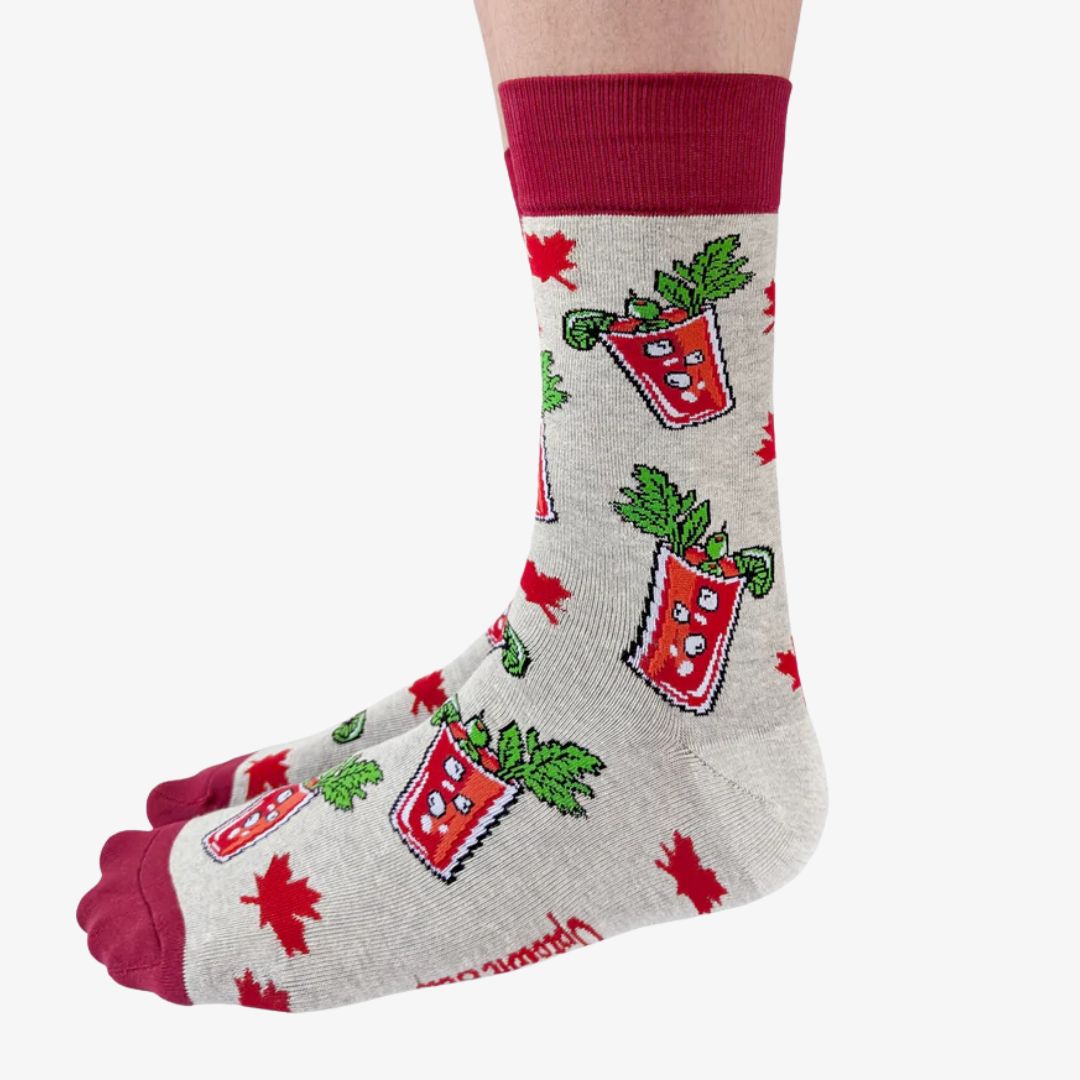 A pair of grey crew socks featuring a pattern of illustrated Canadian Caesar cocktails in red glasses with celery stalks and lime wedges, interspersed with small red maple leaves. The cuff, heel, and toe of the sock are a deep red. The brand "Uptown Sox" is subtly visible on the sole.