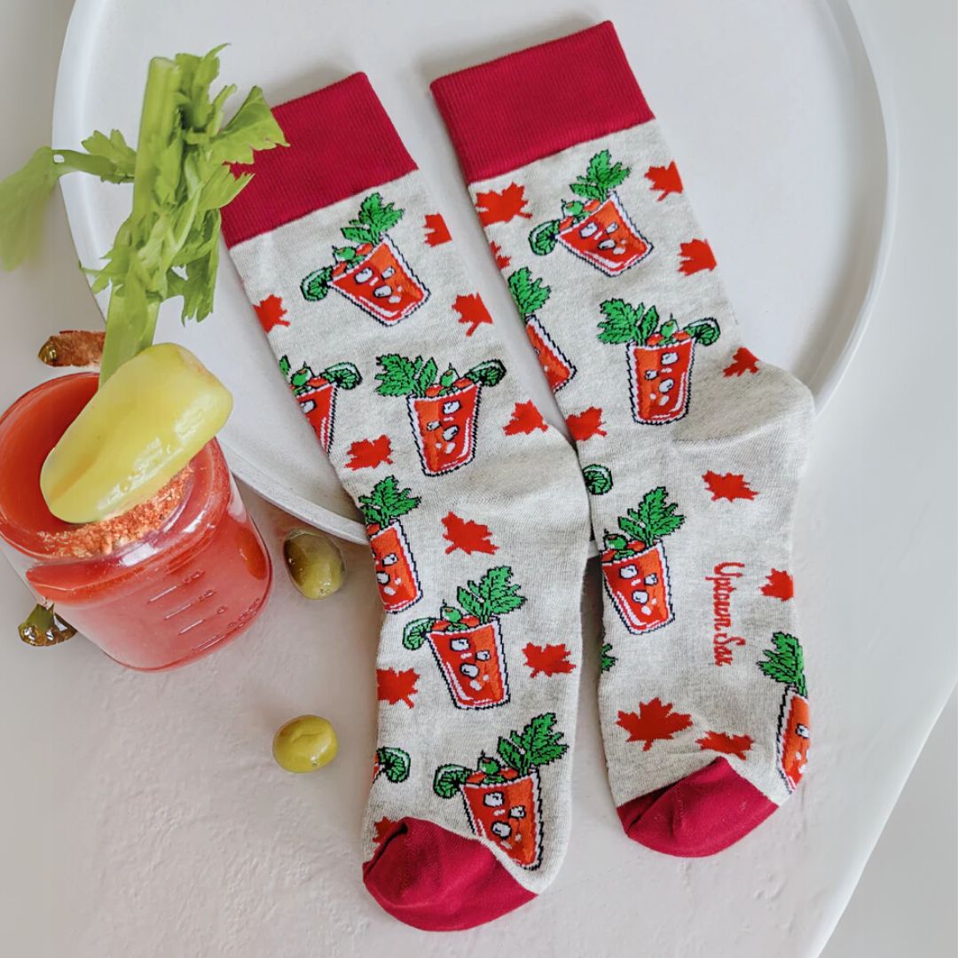 A pair of grey crew socks featuring a pattern of illustrated Canadian Caesar cocktails in red glasses with celery stalks and lime wedges, interspersed with small red maple leaves. The cuff, heel, and toe of the sock are a deep red. The brand "Uptown Sox" is subtly visible on the sole.
