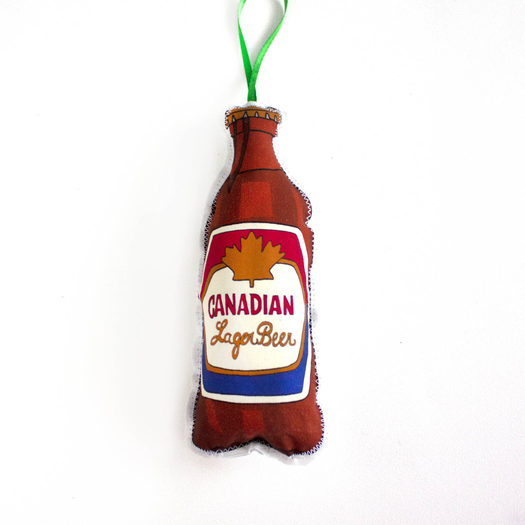 A close-up, high-angle flat lay shot on a plain white background features a soft, plush Christmas ornament shaped like a Canadian beer bottle. The illustration depicts a brown bottle with a label featuring a red maple leaf, with the text "Canadian Lager Beer" in a script font. The ornament is slightly puffy. A bright green ribbon loop is attached to the top for hanging.