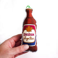 Plush toy shaped like a beer bottle labeled 'Canadian Lager Beer' held by a hand on a white background