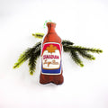 A close-up, high-angle flat lay shot on a plain white background features a soft, plush Christmas ornament shaped like a Canadian beer bottle. The illustration depicts a brown bottle with a label featuring a red maple leaf, with the text "Canadian Lager Beer" in a script font. The ornament is slightly puffy. A bright green ribbon loop is attached to the top for hanging.