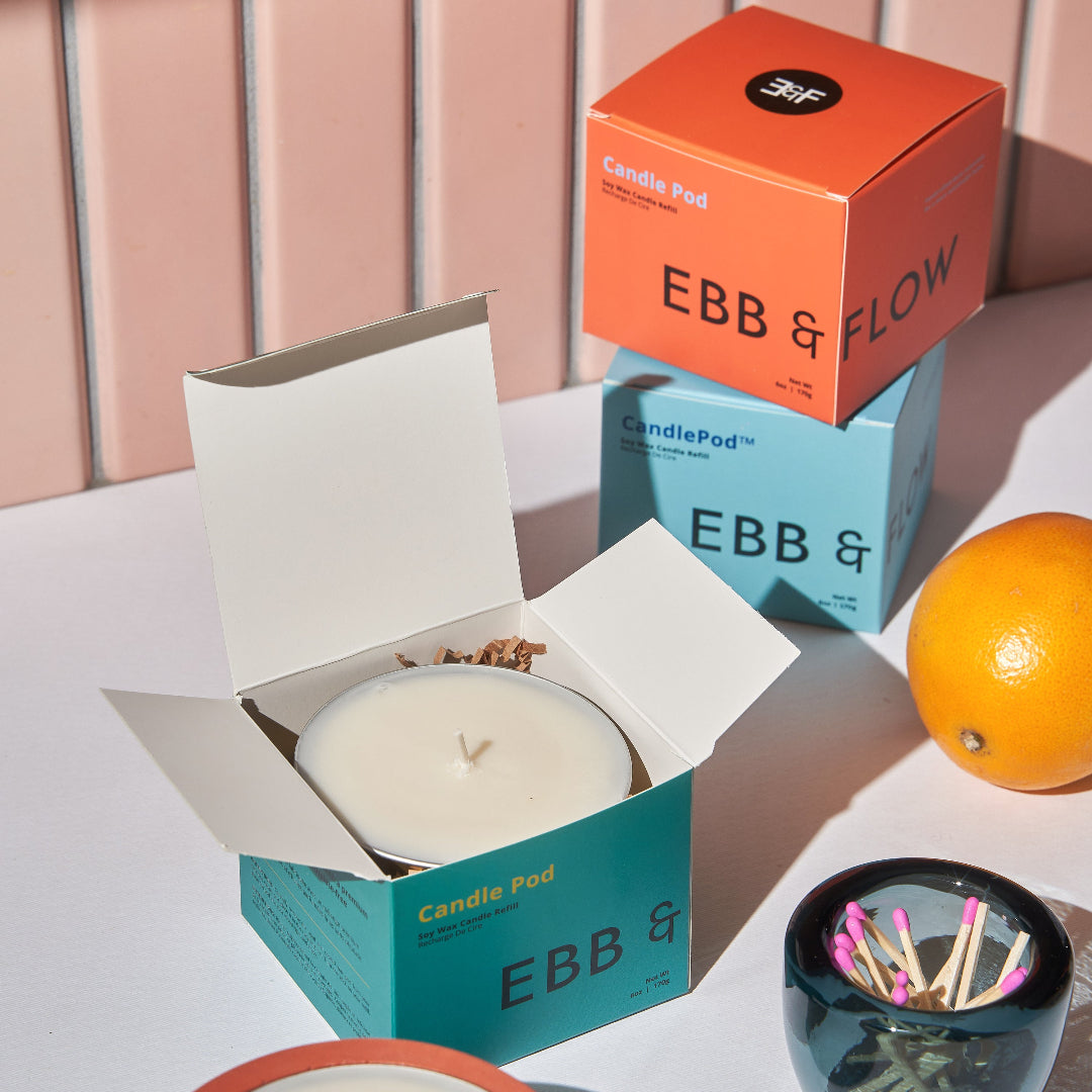 EBB & FLOW CandlePod wax candles shown in their colorful packaging, with one open teal box, one lit candle in an orange container, and another closed orange box stacked on top of a blue box, styled with an orange and a matchbox on a white surface.