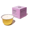 A small lavender scented candle in a gold metal bowl with white wax, shown next to its light purple packaging box on a white background.