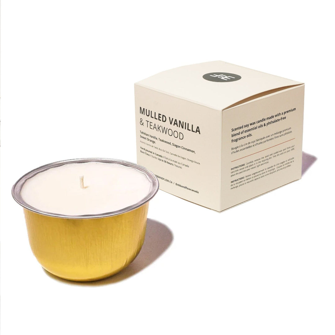 A single-wick candle in a brushed gold metal cup with white wax, shown next to its cream-colored packaging box labeled 'Mulled Vanilla & Teakwood'.