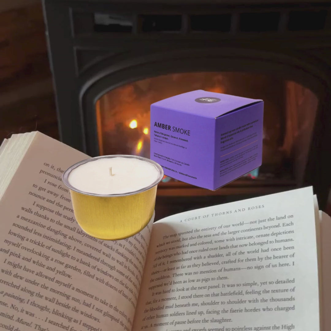 A small amber candle in a white cup sits on an open book in front of a warm fireplace, with its purple box behind it. The candle label reads 'AMBER CANDLE'.