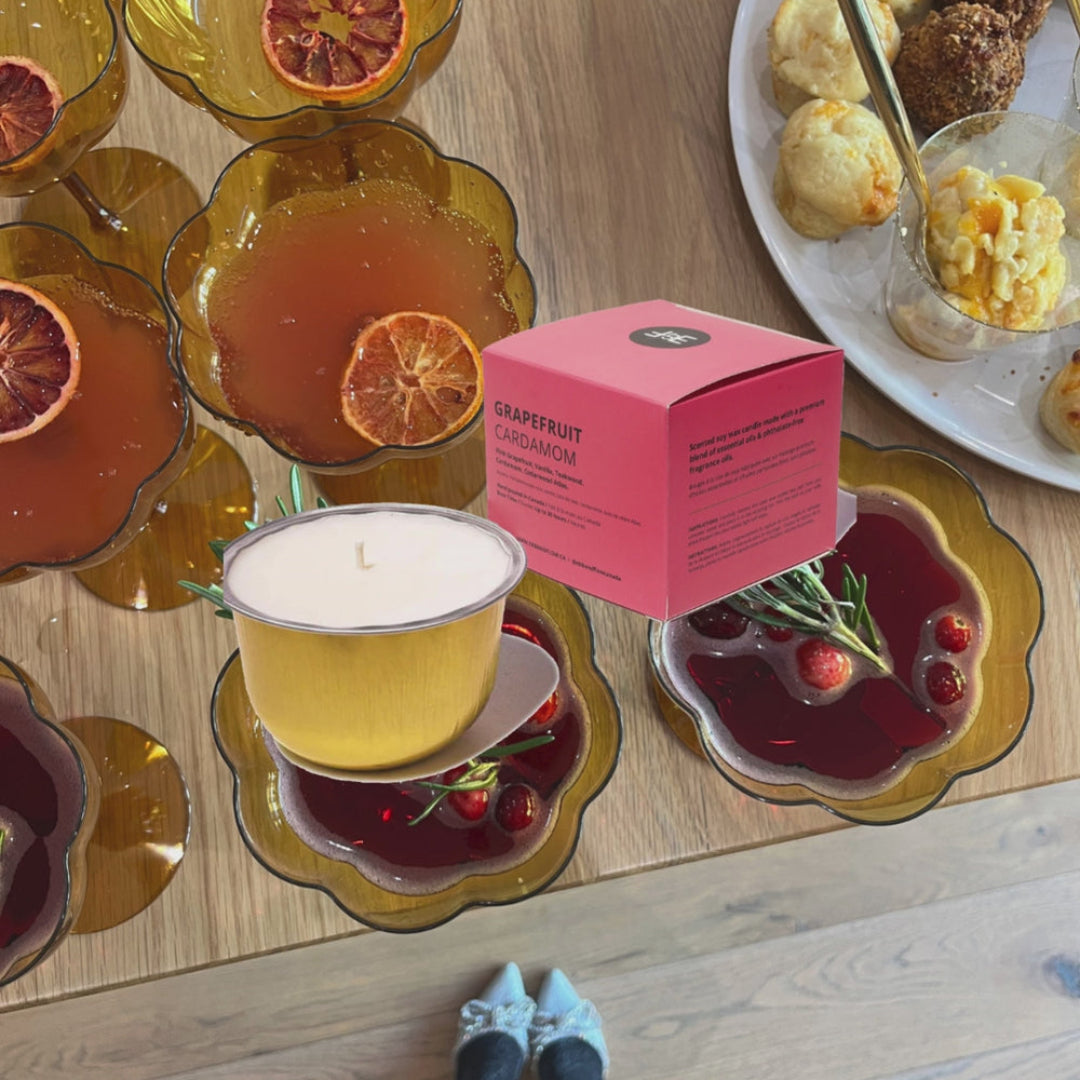 A pink box labeled 'Grapefruit Champagne' candle scent is shown next to a yellow candle in a glass holder on a wooden floor, surrounded by amber glass plates with citrus slices and a plate of pastries.