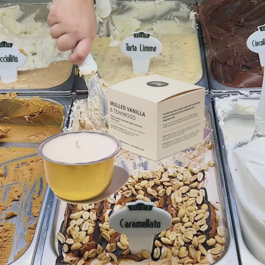 A hand is serving a scoop of vanilla and teamwood-flavored gelato from a metal tray, with a small candle in front of it. The scene shows multiple gelato flavors in a display case, including Caramellato with peanuts and chocolate drizzle, and a box labeled Malloped Vanilla & Teamwood.