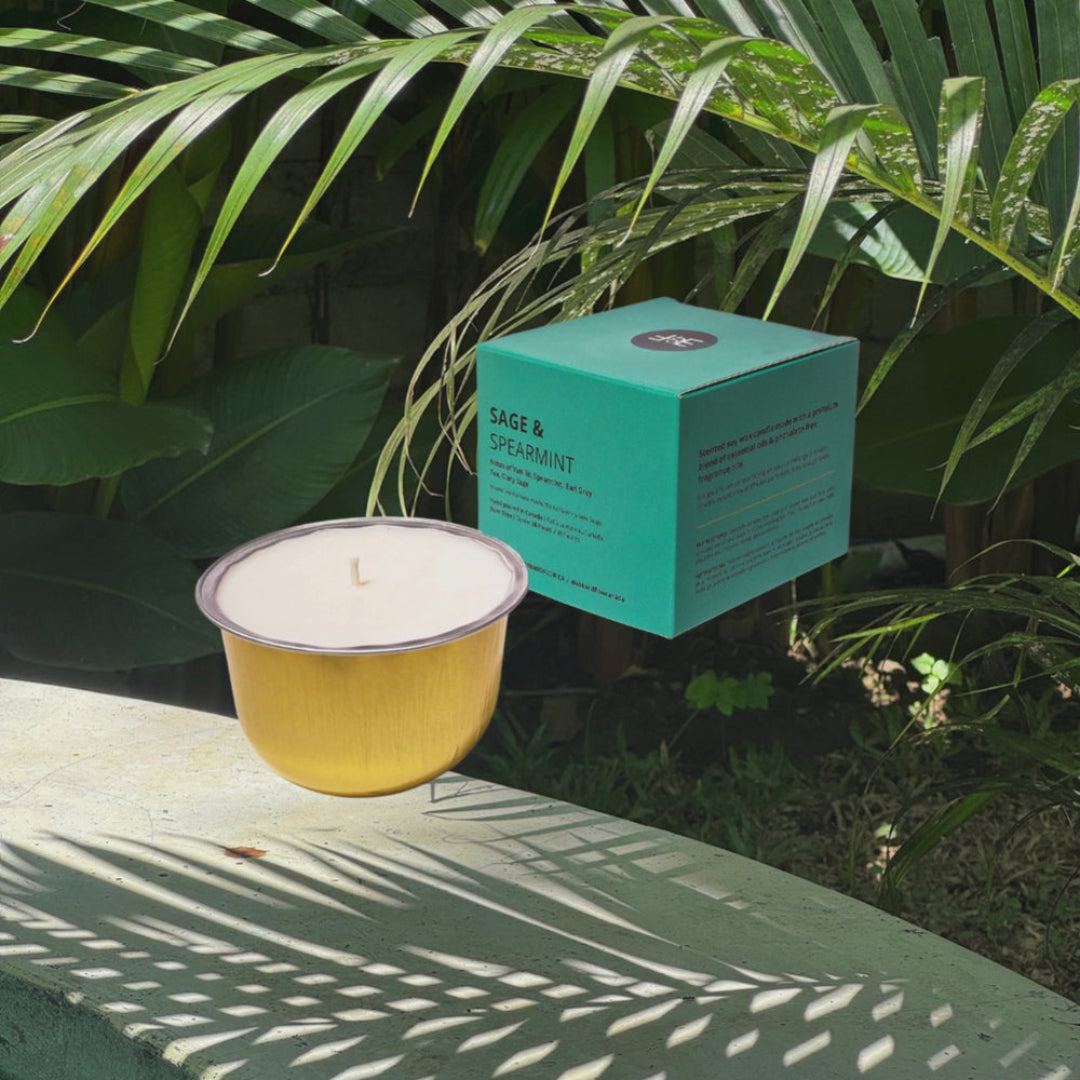 A yellow candle in a metal cup sits beside a turquoise product box labeled Sage & Spearmint, placed outdoors on a stone surface with palm leaves and dappled sunlight in the background.