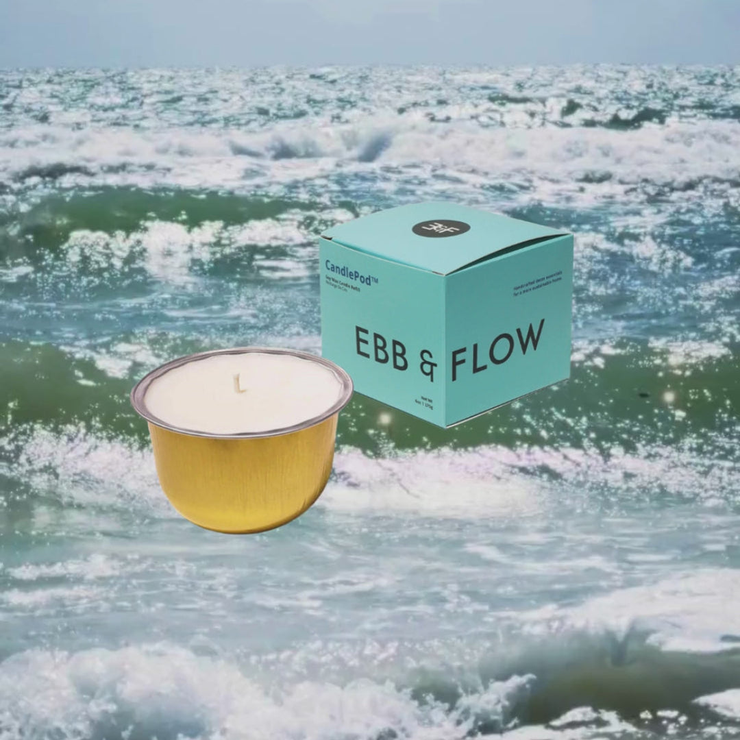 A gold metal candle pot with white wax sits in front of a mint green box labeled EBB & FLOW, set against a background of ocean waves under a pale sky.