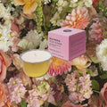 A pink square product box labeled 'WILD ROSE & PEONY FLOWER' sits beside a yellow candle tin with white wax, surrounded by a dense arrangement of pastel flowers.