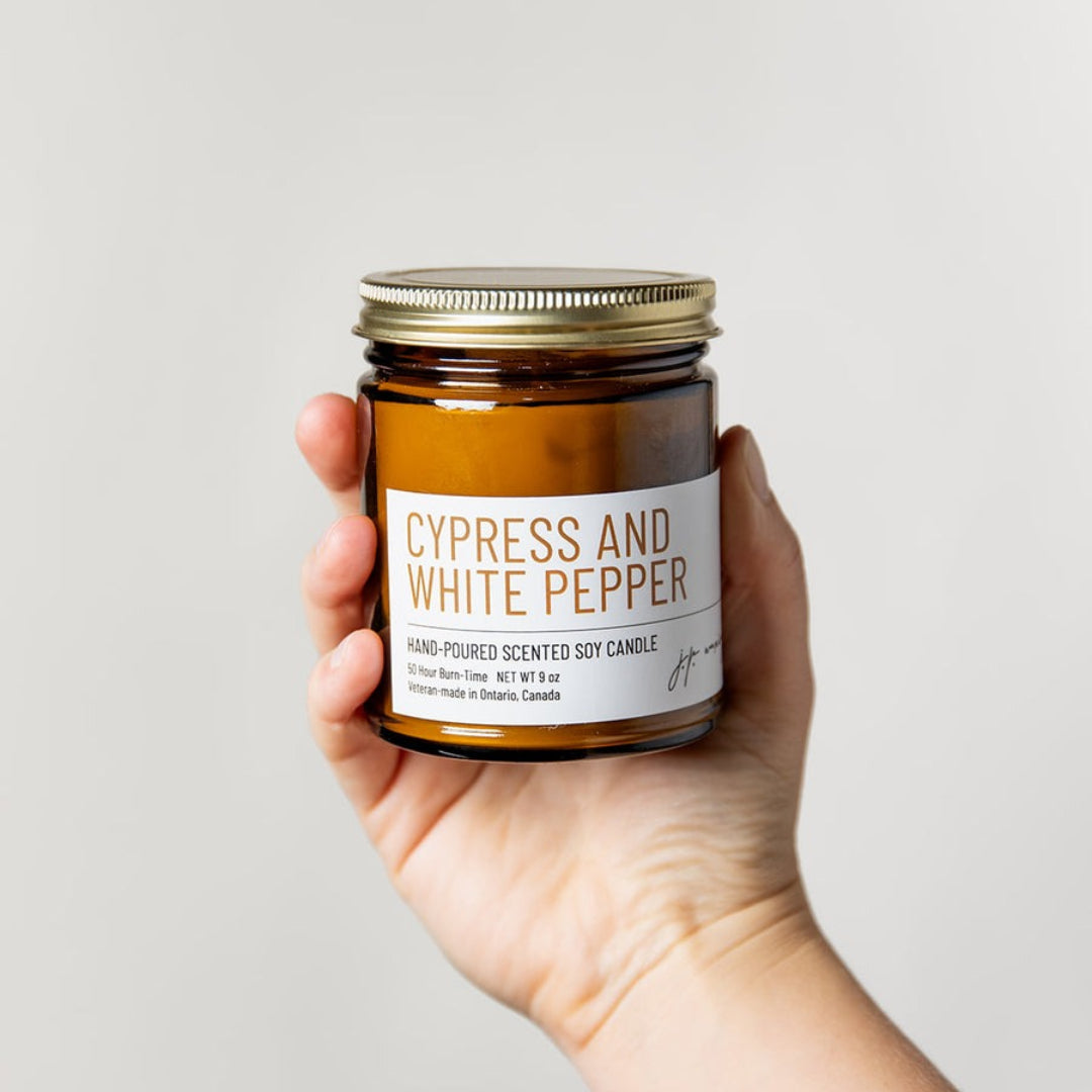 Hand holding a jar of 'Cypress and White Pepper' candle against a plain background
